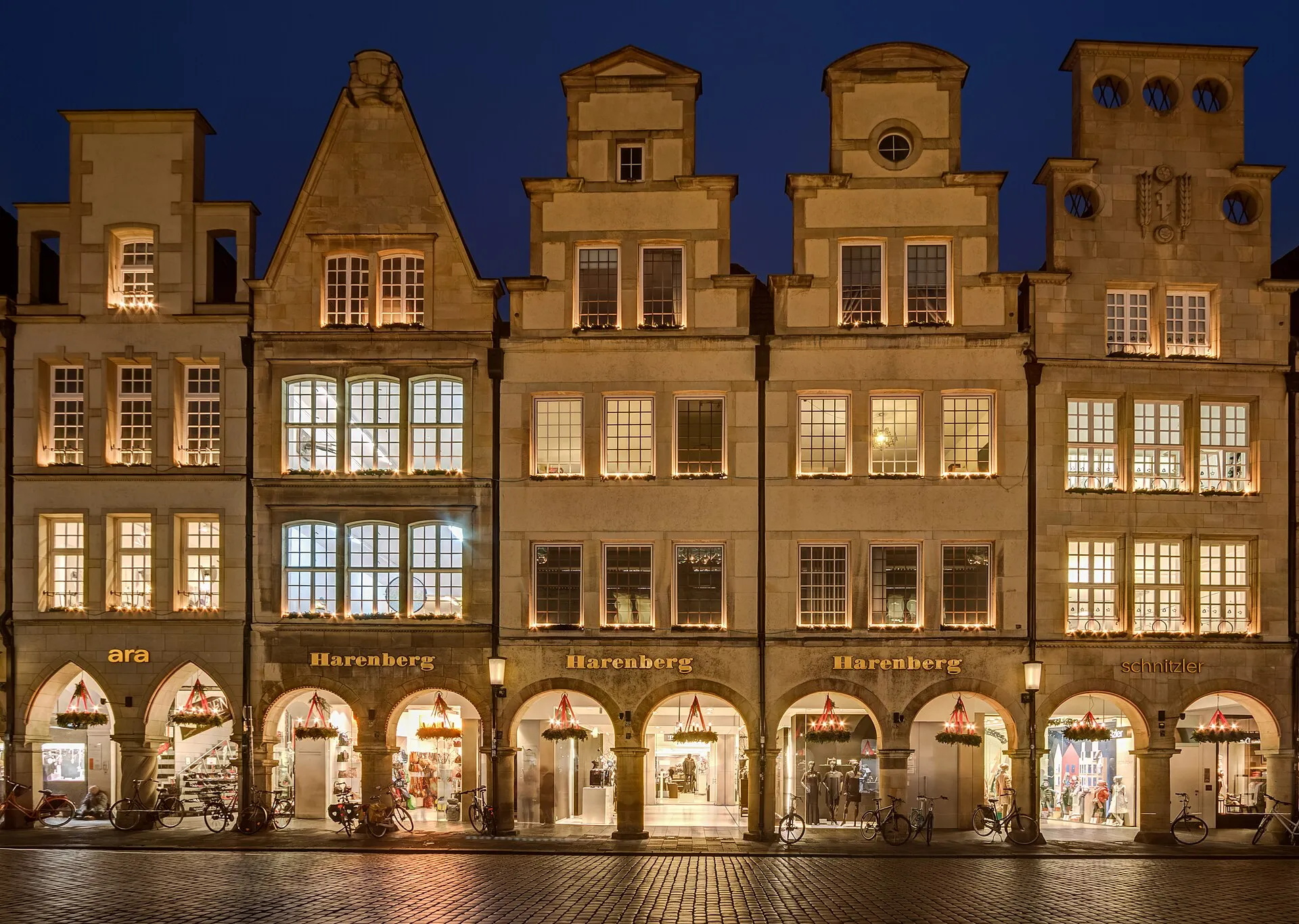 Row of historic stone gabled houses at blue hour with warm light in every window and arcade